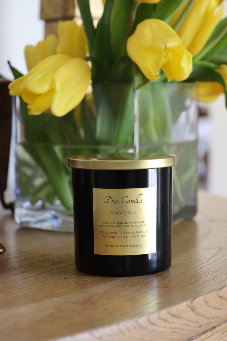 Pu&ograve; includere: Un barattolo di candela nero con un coperchio e un'etichetta dorati. L'etichetta riporta "Dojo Candles" e "Marvelous". La candela &egrave; circondata da tulipani gialli in un vaso di vetro. La candela pesa 255 grammi ed &egrave; realizzata con cera di soia 100% naturale.