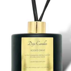 May include: Black glass bottle with a gold lid and black reeds. The bottle has a gold label with the text "Dojo Candles Rodeo Drive Notes Cedarwood, Papyrus, Leather Accord, Violet, Musk Luxury Reed Diffuser Volume Approx. 6.8 fl oz (200 ml)"