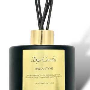 May include: Black glass reed diffuser bottle with a gold lid and black reeds. The label reads "Dojo Candles Ballantyne" and lists the fragrance notes: Bergamot, Petitgrain, Grapefruit, Orange Blossom, Cedar, Amber, Musk and Vetiver. The label also states "Luxury Reed Diffuser" and "Volume Approx. 6.8 fl oz (200 ml)".