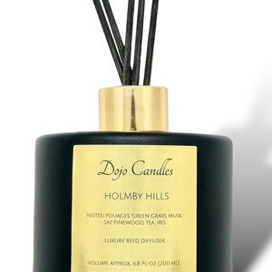 May include: Black glass reed diffuser bottle with a gold lid and black reeds. The label reads "Dojo Candles Holmby Hills" and lists the notes: "Foliages, Green Grass, Musk, Sap Pinewood, Tea, Iris". It also says "Luxury Reed Diffuser" and "Volume Approx. 6.8 fl oz (200 ml)".
