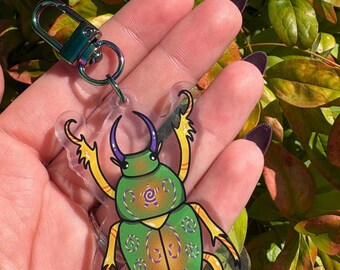 May include: A clear acrylic keychain featuring a vibrant green and yellow beetle design. The beetle has purple horns and intricate patterns on its shell. It is attached to a silver-colored clasp and ring. The background is a blur of green and yellow leaves.