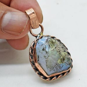 Könnte beinhalten: Kupferfarbener Anhänger mit einem hellblauen und grünen Stein. Der Stein hat eine unregelmäßige Form und ist von einem gedrehten Kupferdraht umrahmt. Der Anhänger hat eine Kupferschlaufe für eine Kette.