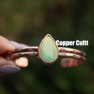Könnte beinhalten: Ein Kupferarmreif mit einem zentralen, tropfenförmigen hellgrünen Stein in einer Zargenfassung. Das Band hat ein zweizeiliges Design mit eingestanzten Sonnenstrahlenmustern auf beiden Seiten. Der Text Copper Cultt ist rechts zu sehen.