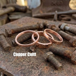 Könnte beinhalten: Drei Kupferringe mit gehämmertem Finish liegen zwischen mehreren rostigen Metallschrauben. Die Ringe unterscheiden sich in Dicke und Oberfläche, wobei einer eine deutlich gehämmerte Struktur aufweist. Der Text Copper Cultt ist zu sehen.