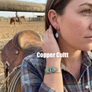 Könnte beinhalten: Ein Kupferarmreif mit zwei herzförmigen Türkissteinen und einem gewellten, strukturierten Band, kombiniert mit einem Ohrhänger in Form eines Halbmonds mit ovalem Stein. Der Text Copper Cultt ist in der Bildmitte zu sehen.