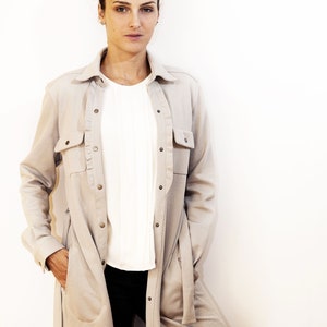 May include: A woman wearing a light beige long-sleeve shirt with two front pockets and a button-down closure. Underneath, she wears a white blouse and black pants. The shirt has a relaxed fit and a collar.