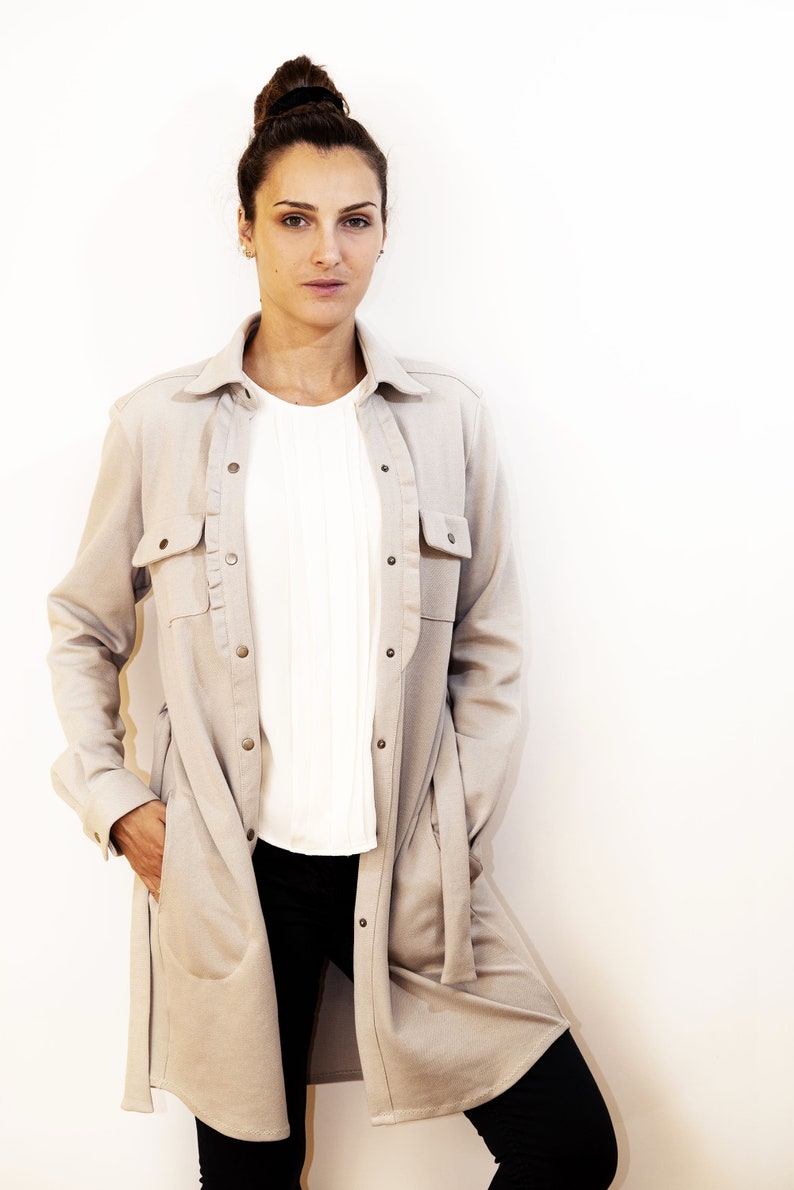 May include: A woman wearing a light beige long-sleeve shirt with two front pockets and a button-down closure. Underneath, she wears a white blouse and black pants. The shirt has a relaxed fit and a collar.