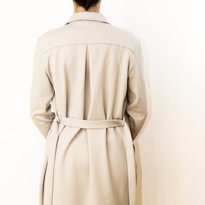 May include: A light beige trench coat with a matching belt. The coat features a classic collar, a pleated back, and long sleeves. The model is wearing black pants. The coat is made of a soft, neutral-toned fabric.