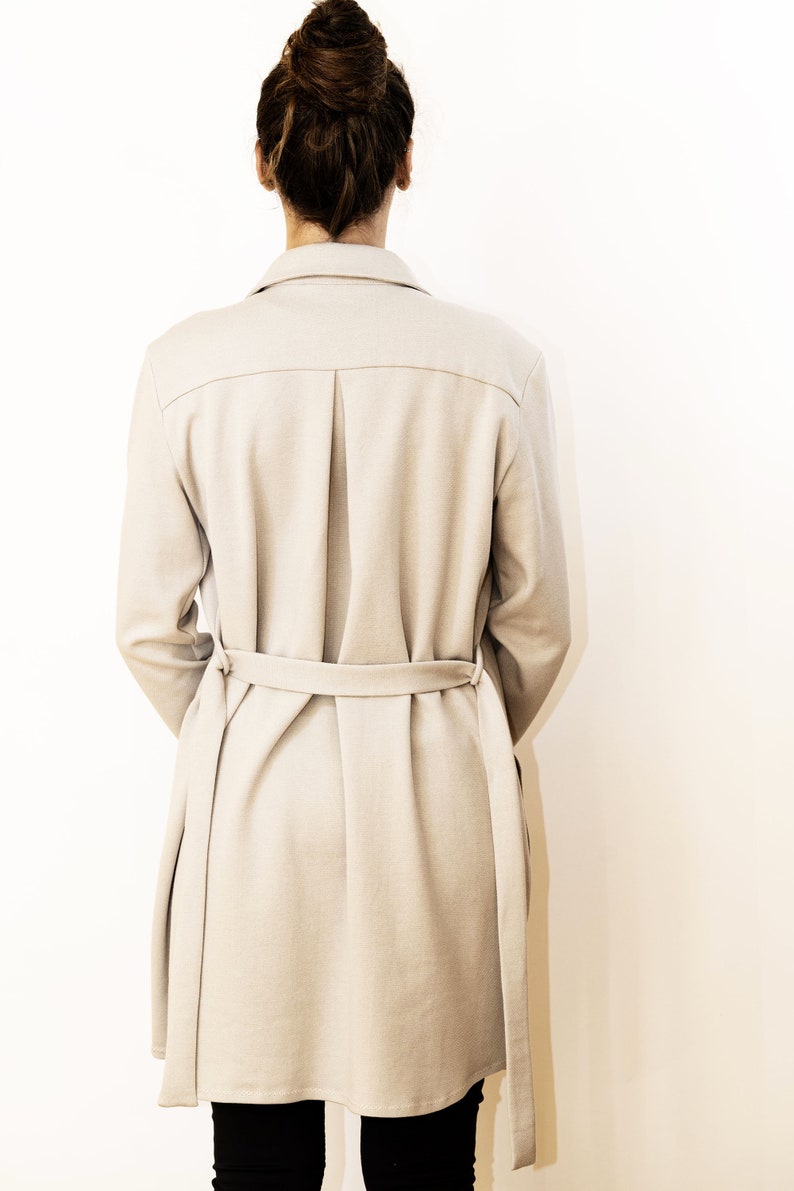 May include: A light beige trench coat with a matching belt. The coat features a classic collar, a pleated back, and long sleeves. The model is wearing black pants. The coat is made of a soft, neutral-toned fabric.