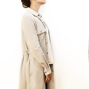 May include: A beige long coat with a matching belt and buttoned pockets. The coat is worn over a white blouse and black pants. The model has her hair up in a bun and is looking up.