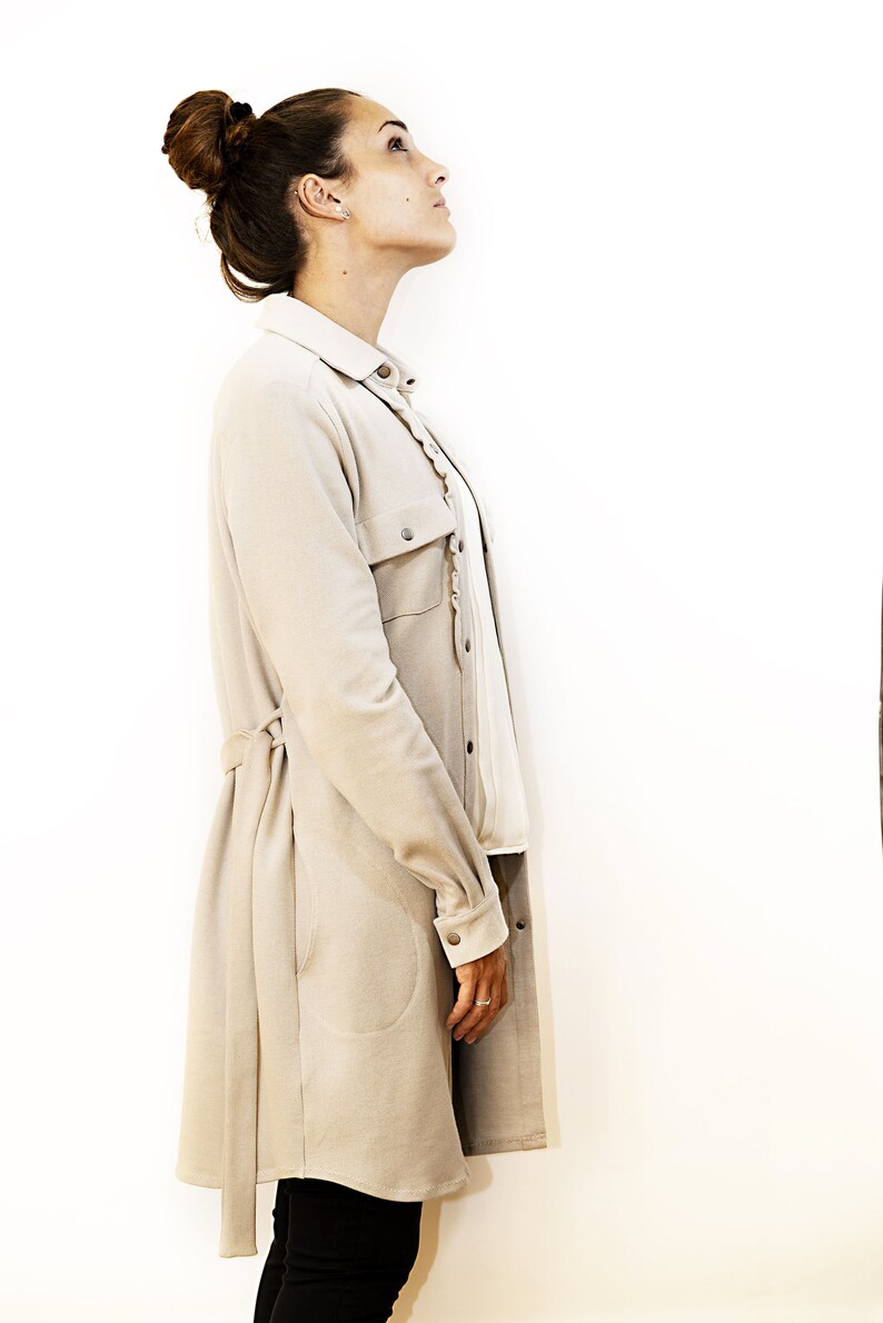 May include: A beige long coat with a matching belt and buttoned pockets. The coat is worn over a white blouse and black pants. The model has her hair up in a bun and is looking up.