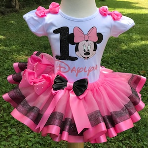 Può includere: Tutu rosa e nero con una maglietta bianca con un numero uno nero e un'immagine di Minnie Mouse. La maglietta ha anche il testo "Daylon".
