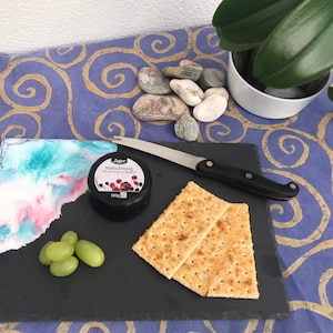 Puede incluir: Una tabla de queso rectangular de pizarra con un diseño abstracto colorido, acompañada de queso Wensleydale, galletas saladas, uvas y un cuchillo para queso. La tabla está sobre una tela azul y dorada estampada.