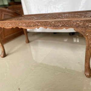 May include: A carved wooden coffee table with intricate floral designs. The table has four legs with a curved design and a rectangular top.