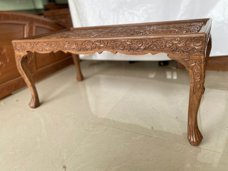 May include: A carved wooden coffee table with intricate floral designs. The table has four legs with a curved design and a rectangular top.