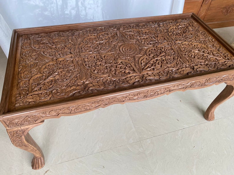 May include: A intricately carved wooden coffee table with a floral design. The table has four legs and a rectangular top.