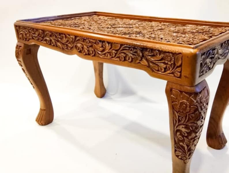 May include: A wooden coffee table with intricate carvings of flowers and leaves. The table has four legs with a curved design and a rectangular top.