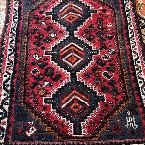 May include: A red, black, and white rug with a geometric pattern. The rug has a fringe border.