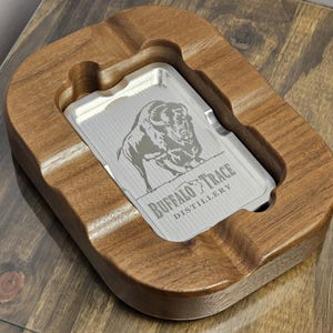 Puede incluir: Un cenicero de madera con un inserto plateado. El inserto presenta un grabado detallado de un bisonte y el texto "Buffalo Trace Distillery". El cenicero es de madera oscura y tiene forma rectangular con bordes redondeados.