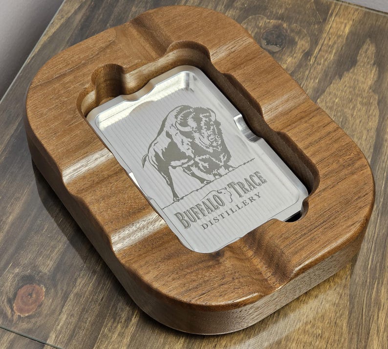 Puede incluir: Un cenicero de madera con un inserto plateado. El inserto presenta un grabado detallado de un bisonte y el texto "Buffalo Trace Distillery". El cenicero es de madera oscura y tiene forma rectangular con bordes redondeados.