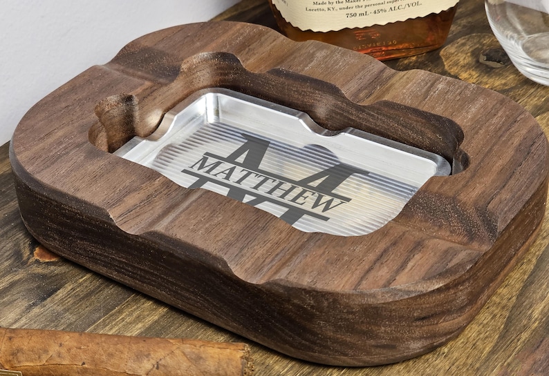 Puede incluir: Un cenicero de madera personalizado con un inserto de metal plateado. El inserto est&aacute; grabado con el nombre "MATTHEW" en una fuente estilizada. El cenicero es de madera oscura y tiene forma rectangular curva.