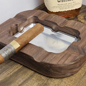 Puede incluir: Un cenicero de madera con un cigarro encima. El cenicero es de madera marr&oacute;n oscuro y tiene un inserto de metal plateado. El cigarro tiene una banda alrededor. El cenicero est&aacute; sobre una superficie de madera.