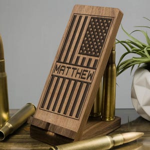 May include: A wooden phone stand with a carved American flag and the name "MATTHEW". The stand is made of dark wood and has a rustic look. It is supported by four gold-colored bullet casings.