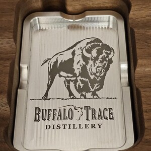 Puede incluir: Una bandeja rectangular plateada con el logotipo de Buffalo Trace Distillery y un grabado de bisonte, colocada en un soporte de madera. El texto en la parte superior dice "Super fine detailed engraving!" y en la inferior "Any logo or design you want!"
