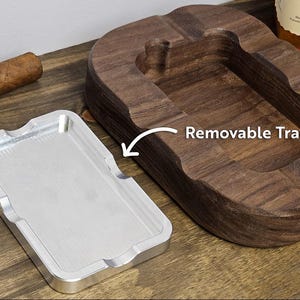 Puede incluir: Un cenicero de cigarros de madera con una bandeja plateada extra&iacute;ble. El cenicero es de madera oscura y tiene un dise&ntilde;o curvo. Un cigarro descansa sobre la superficie de madera. El texto "Removable Tray" es visible.