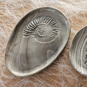 Könnte beinhalten: Zwei graue Keramik-Ringschalen. Eine ist oval mit einem spiralförmigen Fossilienmuster. Die andere ist rund mit einem Marmormuster und zwei Ringen. Die Schalen stehen auf einer strukturierten, hellbraunen Oberfläche.
