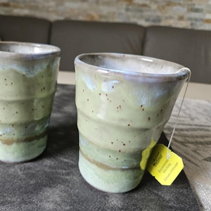 Könnte beinhalten: Zwei handgefertigte Keramikbecher mit gerippter, konischer Form. Die Glasur zeigt eine hellgrüne Basis mit braunen Sprenkeln, die zu einem weißen Rand übergeht. Ein gelbes Teebeutel-Etikett mit der Aufschrift Meßmer ist am rechten Becher zu sehen.