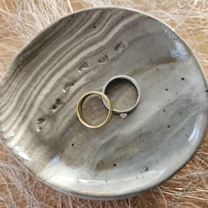 Könnte beinhalten: Ein goldener Ring und ein silberner Ring mit einem kleinen Diamanten liegen auf einer grau-weißen Keramikschale. Die Schale hat ein marmoriertes Design und eine unregelmäßige Form mit kleinen Vertiefungen am Rand.