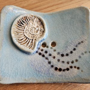 Könnte beinhalten: Hellblaues Keramik-Seifenschale mit einem erhabenen, cremefarbenen Ammoniten-Fossil-Design. Die Schale hat eine Craquelé-Glasur und zwei Abflusslöcher. Schwarze Punktmuster verzieren die Oberfläche. Ein einzigartiges Dekorationsstück.