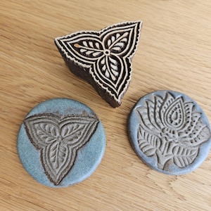 Könnte beinhalten: Ein dreieckiger Holzstempel mit geschnitztem Blattmuster neben zwei runden blauen Keramikfliesen. Eine Fliese zeigt das passende Blattdesign, während die andere ein detailliertes Blumenmotiv mit strukturierten, erhabenen Relieflinien aufweist.