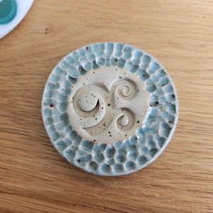 Könnte beinhalten: Ein runder Keramikuntersetzer mit hellblau strukturiertem Rand und cremefarbenem Zentrum. Das Zentrum hat ein Spiraldesign. Der Untersetzer liegt auf einer Holzoberfläche. Im Hintergrund befindet sich ein weißes und türkisfarbenes Objekt.