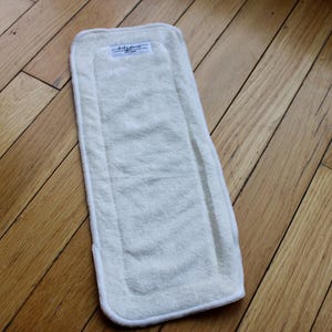 May include: A rectangular, off-white cloth diaper insert with a white border, lying on a wooden floor. The insert has a textured surface and a small label at the top.