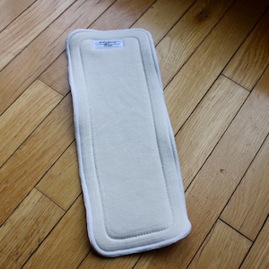 Könnte beinhalten: Ein rechteckiger, cremefarbener Stoffwindeleinsatz mit weißem Rand und Nähten. Der Einsatz liegt auf einem Holzboden. Oben auf dem Einsatz ist ein kleines Etikett zu sehen.