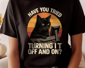 Puede incluir: Camiseta negra con un diseño retro que presenta un gato negro frente a un ordenador. El texto dice "HAVE YOU TRIED TURNING IT OFF AND ON?" en un diseño circular con tonos naranja y amarillo.
