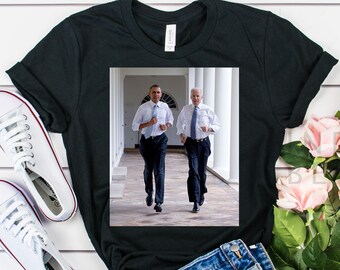 May include: Black t-shirt with a photo of two men in suits running down a hallway. The men are wearing dark suits and white shirts. The hallway is white with columns on either side.