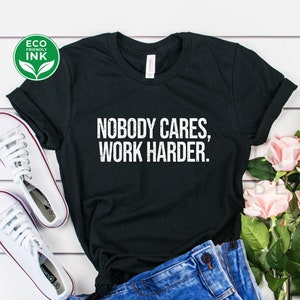 May include: Black t-shirt with the text "Nobody Cares, Work Harder." printed in white. The shirt is displayed on a white wooden surface with a pair of white sneakers, blue jeans, and pink flowers.