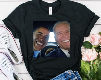 May include: Black t-shirt with a photo of two men smiling at the camera. The men are wearing suits and are in a car. The t-shirt is on a white wooden surface with a pair of white sneakers and a pair of denim shorts.