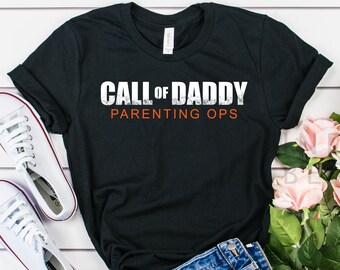 May include: Black t-shirt with white text that reads "Call of Daddy Parenting Ops". The shirt is laid out on a white wooden surface with a pair of white sneakers, denim shorts, and pink flowers.