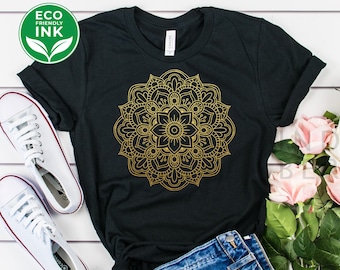 May include: A black t-shirt with a gold mandala design. The shirt is laid out on a white wooden surface with a pair of white sneakers, denim shorts, and a bouquet of pink roses.