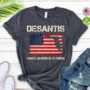 May include: A dark gray t-shirt with a distressed white graphic that says "DESANTIS" with an American flag and the state of Florida superimposed on the flag. The text below the flag says "MAKE AMERICA FLORIDA".