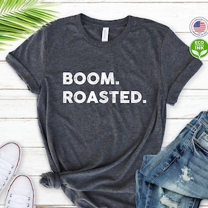 May include: A dark heather grey t-shirt with the text "BOOM. ROASTED." printed in white. The shirt is laid flat on a white wooden surface with a pair of white sneakers and a pair of blue jeans.