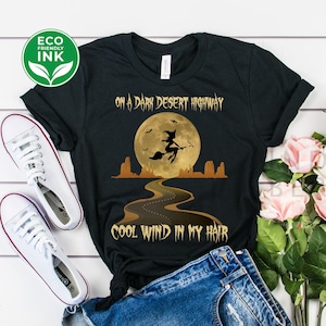 May include: Black t-shirt with the text "ON A DARK DESERT HIGHWAY COOL WIND IN MY HAIR" and a graphic of a witch flying in front of a full moon. The shirt is paired with denim shorts and white sneakers. A bouquet of pink roses is also present.