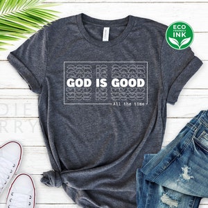 May include: Dark gray t-shirt with the text "GOD IS GOOD" in a white, geometric design. Below the text is "All the time." The shirt is paired with blue jeans and white sneakers, suggesting casual wear. An "Eco Friendly Ink" logo is visible.