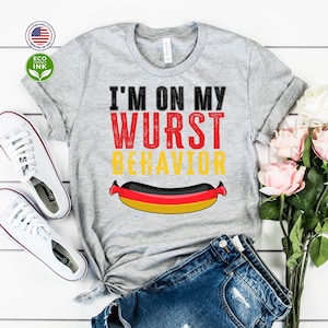 Può includere: Maglietta grigia con il testo "I'm on my Wurst Behavior" in rosso e giallo. Sotto il testo c'è un cartone animato di un hot dog con i colori della bandiera tedesca.