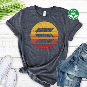 May include: A dark heather grey t-shirt with a vintage-style sunset graphic featuring a stylized letter "S" in the center. The graphic is in shades of yellow, orange, and red.