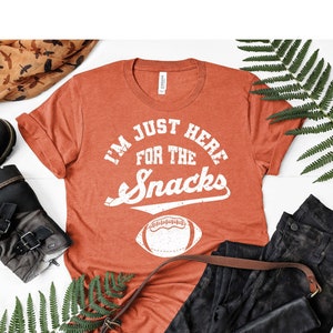 Może przedstawiać: Koszulka w kolorze rdzy z napisem "I'm Just Here For The Snacks" w kolorze białym z białym nadrukiem piłki nożnej.